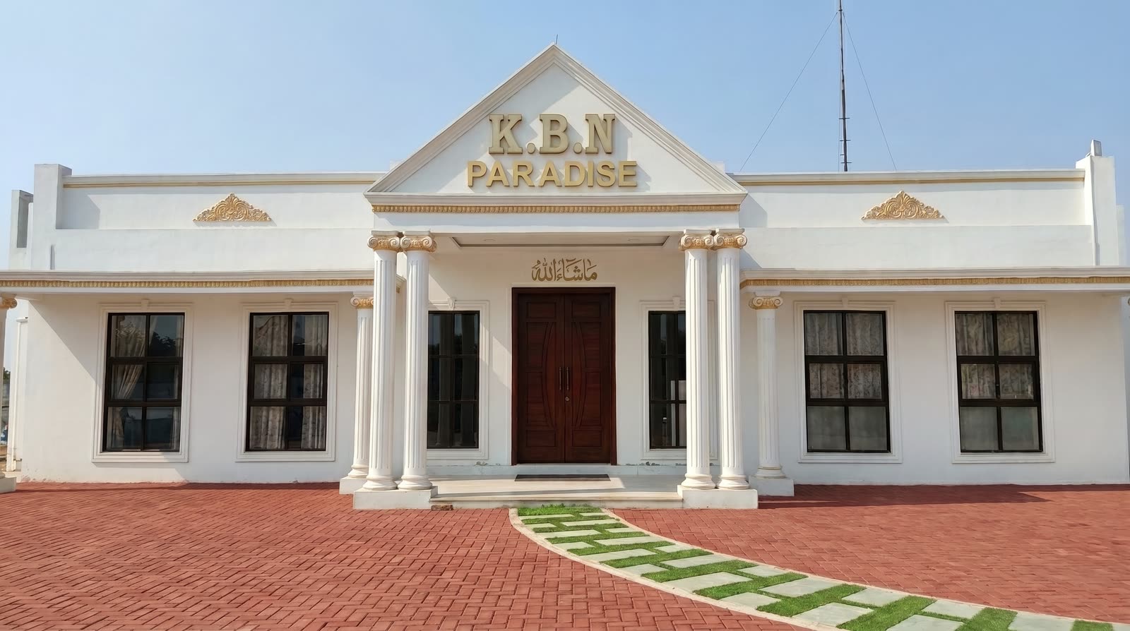 The K.B.N Paradise farmhouse: a white single-storey villa with Ionic columns and the name lettered in gold across the pediment, standing behind a red brick-paved courtyard under a clear sky.