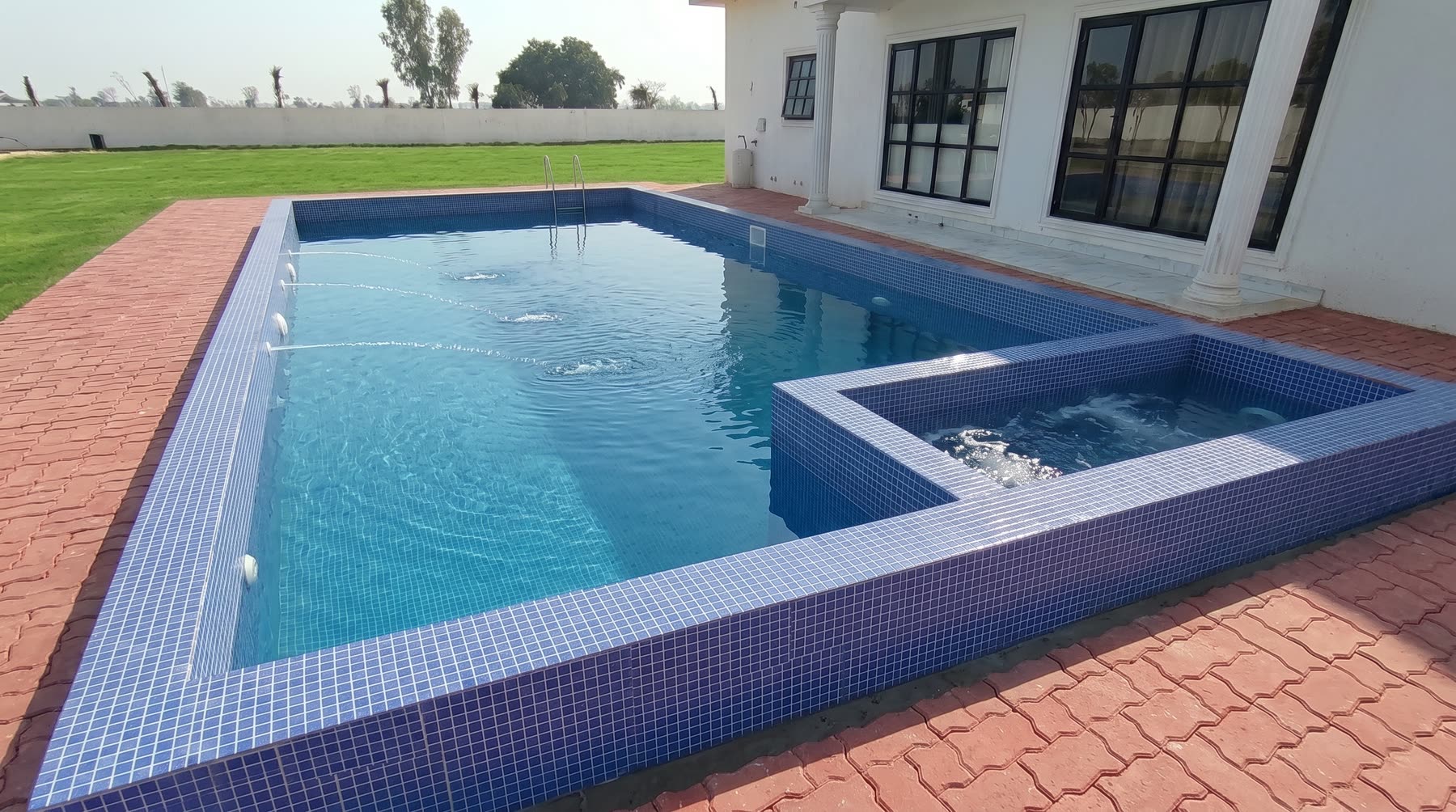 The swimming pool at K.B.N Paradise, tiled in deep blue mosaic, with a raised shallow bathing corner for children at one end, bordered by red brick paving and open lawn.